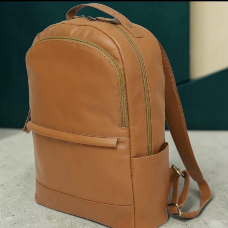 Tan full-grain leather backpack with brass zip closure and padded laptop compartment, shown from the side against a concrete studio backdrop, styled for work, travel, or everyday carry.